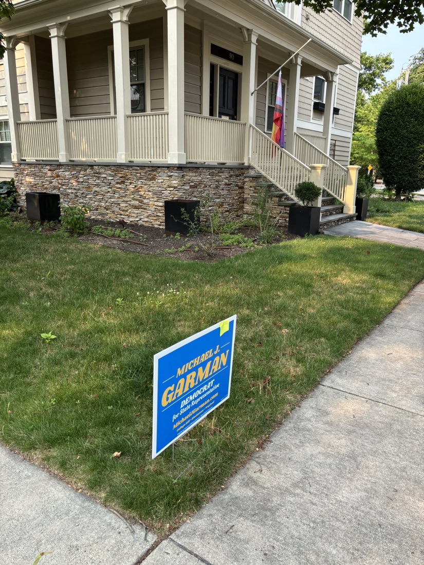 A blue and gold Michael J. Garman yard sign on a College Hill lawn, with a Pride flag flying on the porch behind it